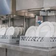Vita tallrikar i diskbackar under en industriell diskmaskin i ett storkök.