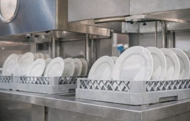 Vita tallrikar i diskbackar under en industriell diskmaskin i ett storkök.